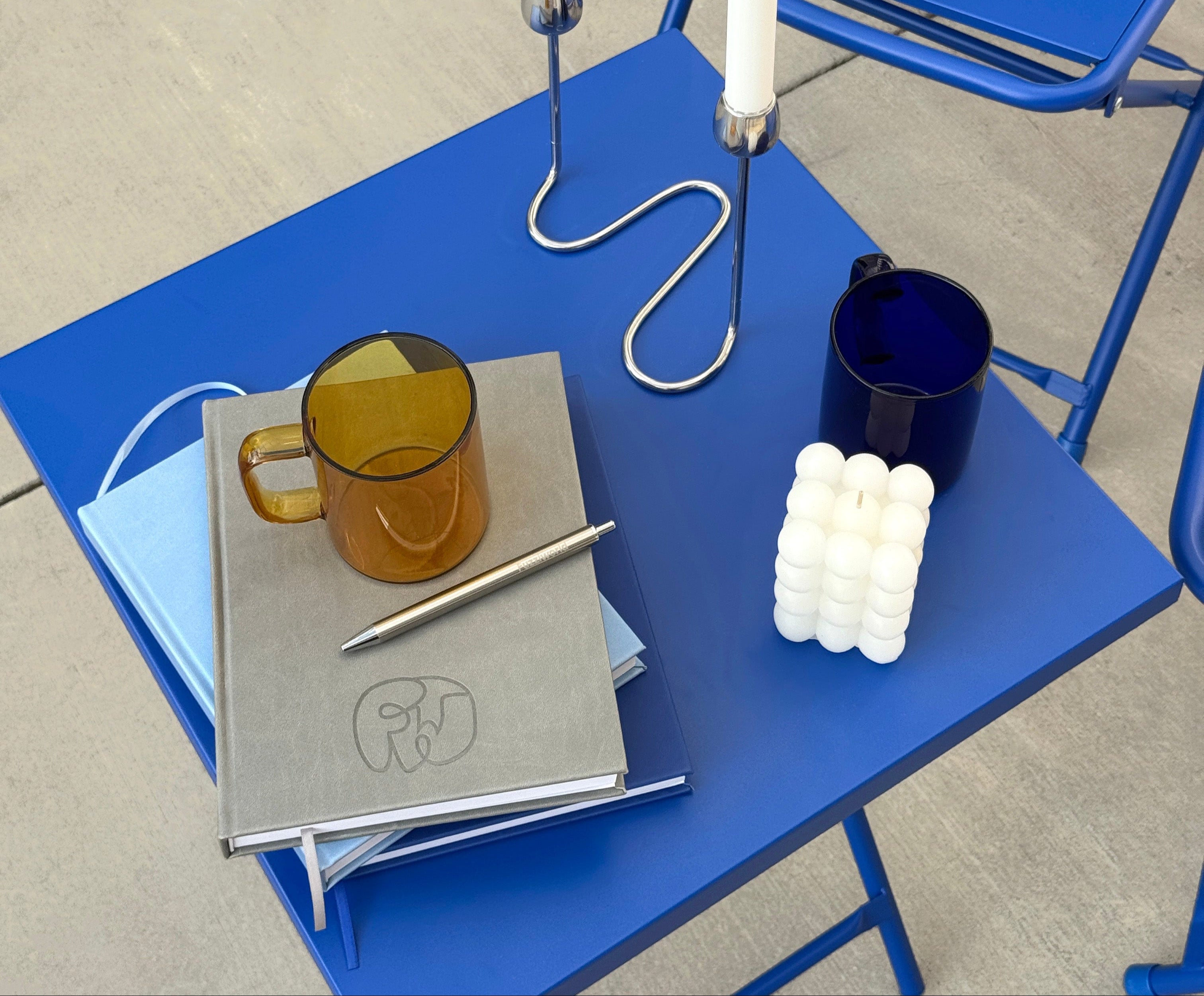 Blue table with a Fuzziworld Journal Notebook, mug, candles, and other tasteful modern decor in an outdoor setting on concrete